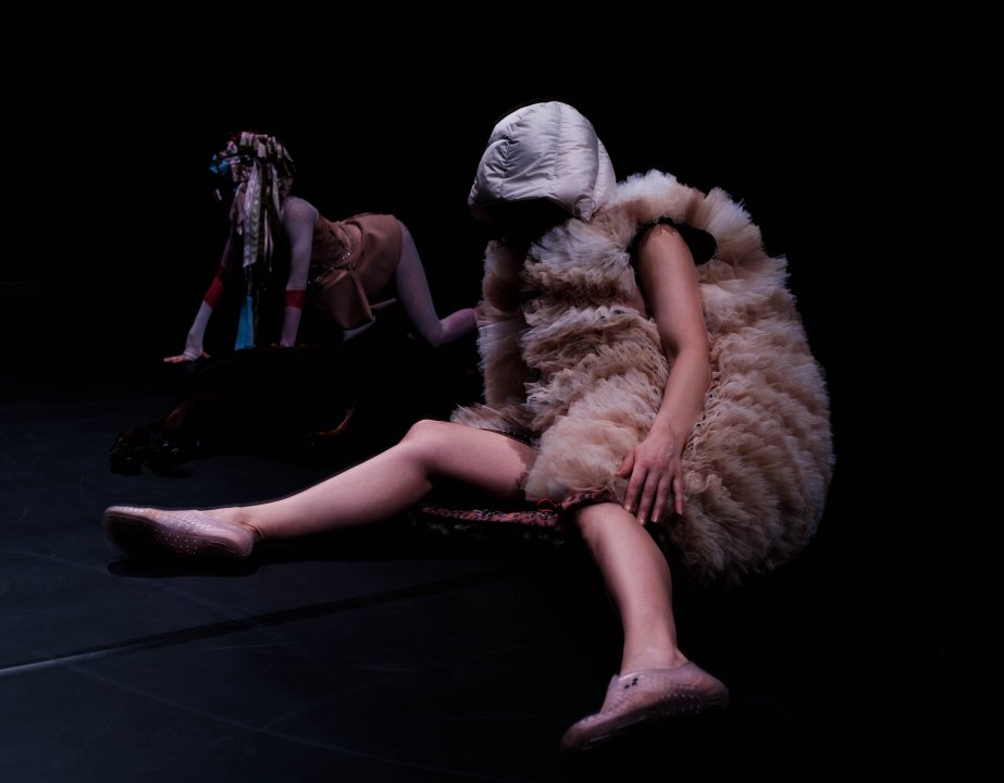 Two people in a dark space, one is on all fours in the back with a layered textile costume on, one is in the front sitting with a tulle costume and bare legs